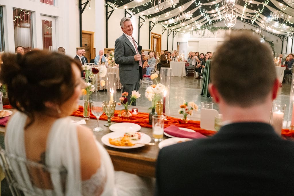 bride and groom look to father of bride while he gives toast